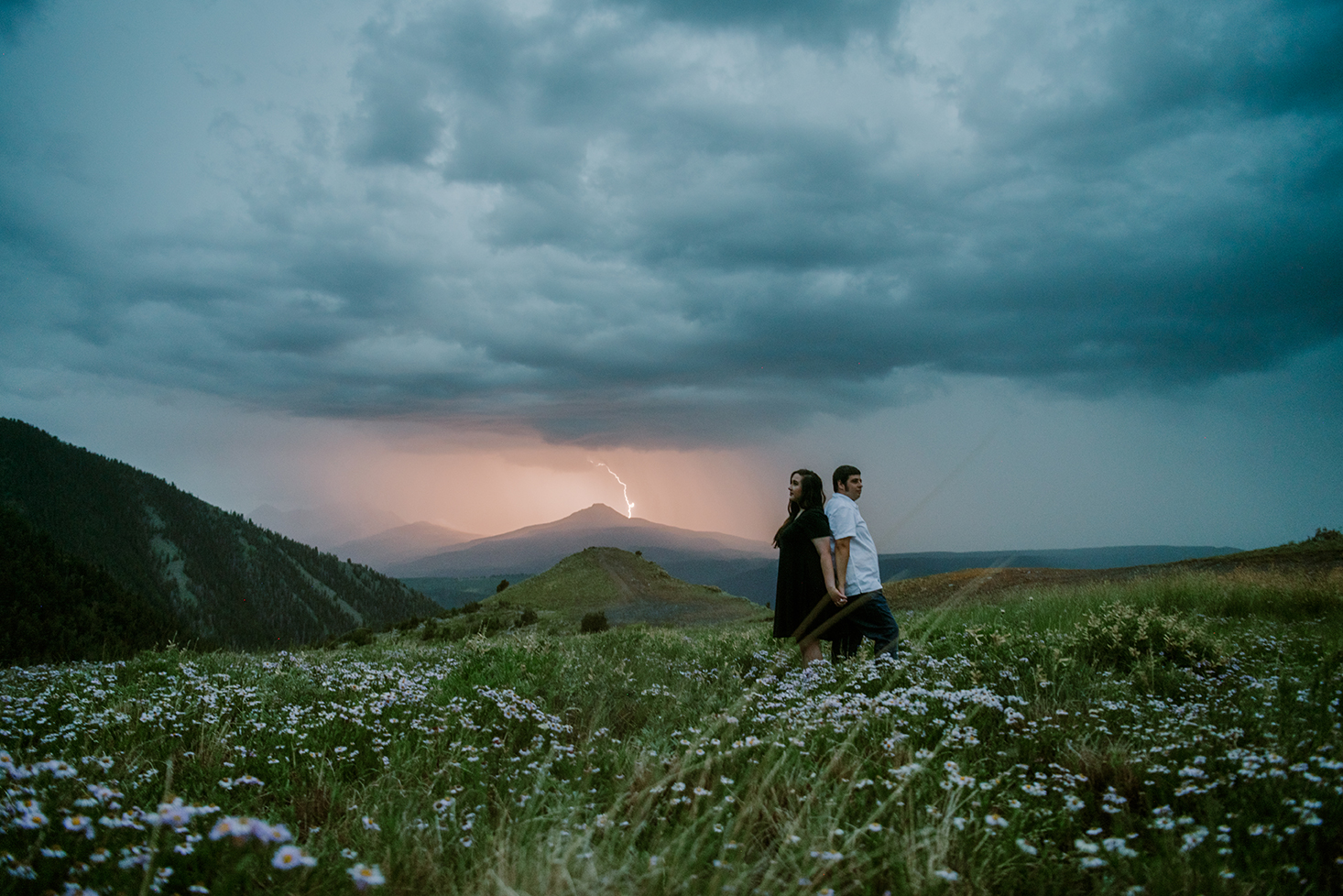 Couple standing in field of flowers as lightning strikes a mountain in the distance